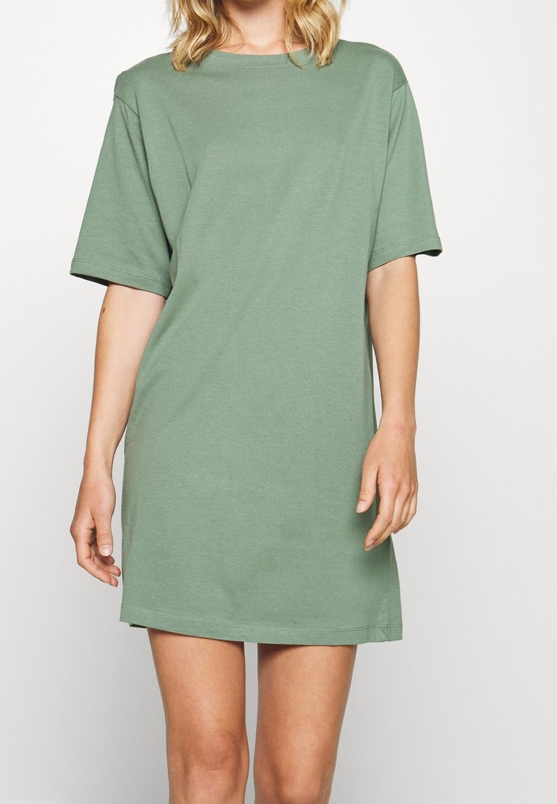 Femme portant une robe T-shirt ample vert sauge à manches courtes, debout devant un fond blanc uni.