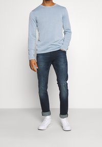 Himmelblaues Langarmhemd kombiniert mit dunklen Jeans und weißen Sneakers. Das Hemd hat einen runden Ausschnitt und eine subtile Textur. Die Jeans weisen abgenutzte Details auf.