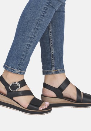 Pieds portant des sandales compensées en cuir noir avec boucle argentée, associées à un jean en denim bleu ajusté.