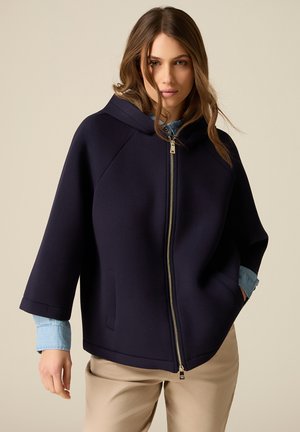 Kobieta ubrana w granatową kurtkę z kapturem zapinaną na zamek z przodu, z widocznymi jasnoniebieskimi rękawami koszuli i beżowymi spodniami, stojąca na jednolitym tle.