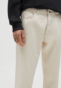 Béžové džínové kalhoty s úzkým střihem a jemnou strukturou. Boční kapsy, kontrastní švy a zapínání na knoflík. Ruka opřená o bok.
