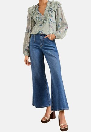 Vrouw draagt een bloemrijke blouse met ruches en lange opgepofte mouwen, blauwe jeans met wijd uitlopende pijpen en beige plateauhakken met enkelbandjes.