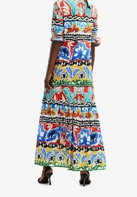 Femme portant une longue robe colorée à motif floral avec les manches retroussées et des sandales noires à talons hauts, debout sur un fond blanc.