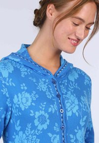 Blau gemusterte Strickjacke mit Kapuze, ausgestattet mit kontrastierenden dunkelblauen Knöpfen und einem strukturierten Strickstoff. Leichtgewichtig mit einer weichen Haptik.