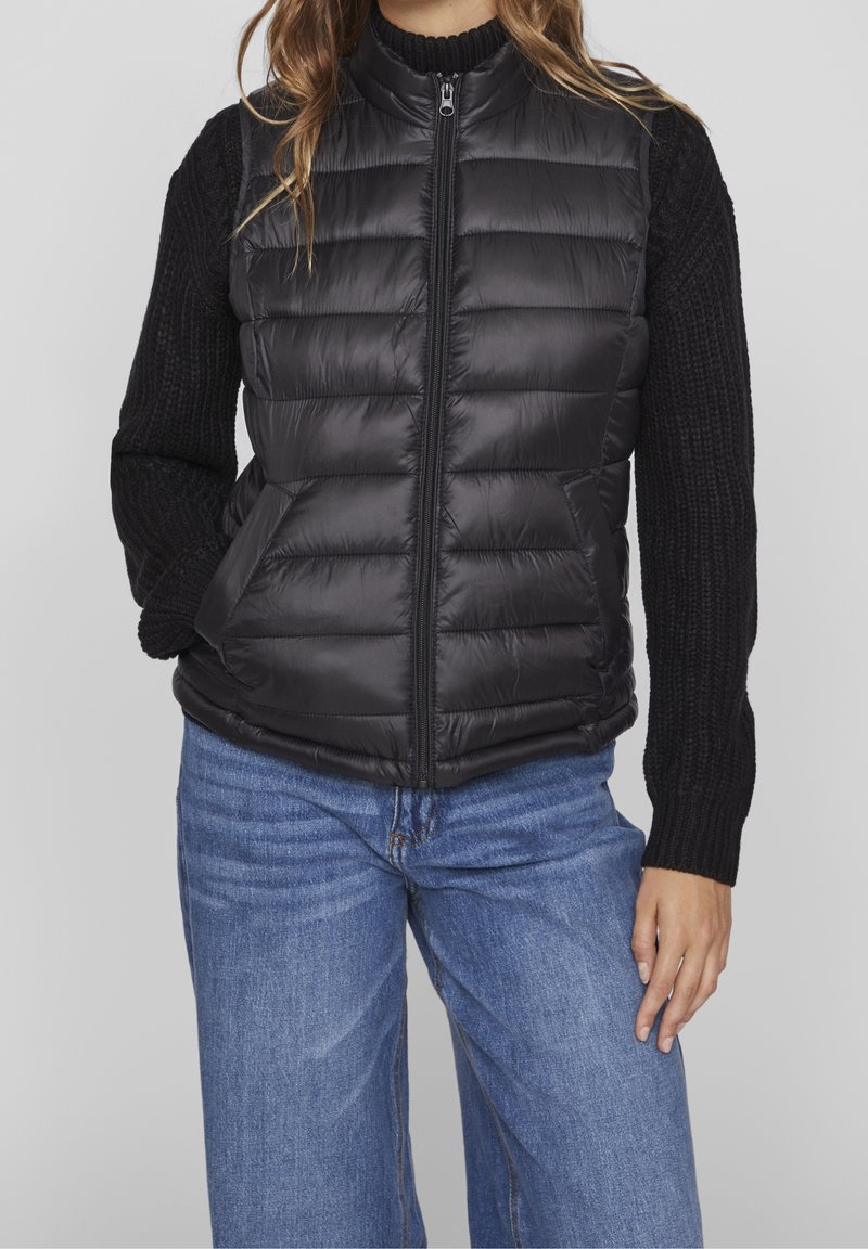 Personne portant un gilet matelassé noir zippé sur un pull en maille noir avec un jean bleu.