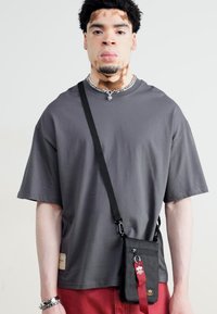 T-shirt grigia oversize, maniche corte, abbinata a una borsa a tracolla nera con dettagli rossi e una tracolla, indossata su pantaloni rossi.
