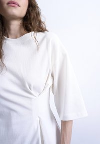 Γυναίκα με κυματιστά καστανά μαλλιά που φοράει ένα λευκό, φαρδύ φόρεμα με μανίκια μέχρι τον αγκώνα και πιέτες κοντά στη μέση.