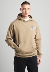Tan hoodie made of cotton blend with kangaroo pocket, featuring "USA New York City" embroidered in blue. Slim fit with adjustable hood.