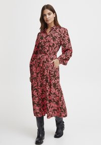 Rochie lungă cu un model floral în nuanțe de roz și maro, având un guler, mâneci lungi, talie strânsă și buzunare laterale. Asortată cu cizme negre.