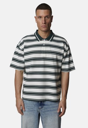 Homme aux cheveux courts portant un polo blanc avec des rayures horizontales foncées et un jean bleu, debout devant un fond uni.