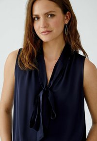 Marineblaue ärmellose Bluse mit einem Krawatten-Halsausschnitt, aus glattem Stoff gefertigt. Verfügt über einen subtilen Glanz und ein minimalistisches Design, das die Einfachheit betont.