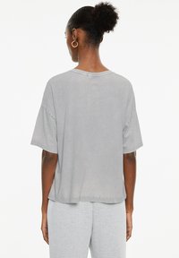 Camiseta de manga corta gris con un corte relajado, cuello redondo y tela de algodón suave. Presenta un dobladillo ligeramente desigual y una textura sutil.