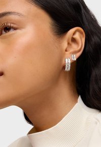 Boucles d'oreilles en argent avec un design géométrique présentant des découpes, portées sur l'oreille d'un modèle sur un fond neutre. Texture lisse.