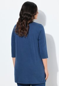 Frau mit langen, welligen dunklen Haaren, die ein blaues Tunika-Oberteil mit drei Viertelärmeln trägt, von hinten vor einem weißen Hintergrund betrachtet.