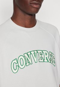 Jasnoszara bluza z zielonym haftowanym napisem "CONVERSE". Posiada okrągły dekolt i rękawy raglanowe, ukazując miękką, gładką teksturę materiału.