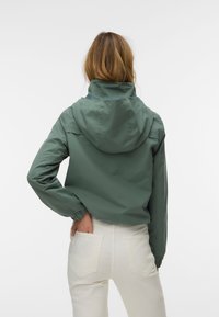 Femme aux cheveux châtain clair portant une veste à capuche verte et un pantalon blanc, debout, une main dans sa poche arrière, de dos.