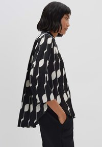 Camicia bianca e nera con righe verticali e motivi circolari, vestibilità ampia, maniche a tre quarti, tessuto morbido, leggero dettaglio di plissettatura.