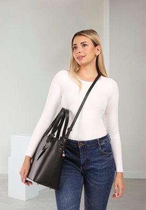Woman wearing white long-sleeve top and blue jeans carrying large black handbag with shoulder strap in bright indoor setting.