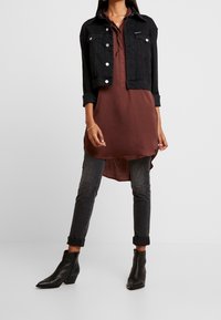 Femme portant une veste en denim noire courte sur une tunique marron, associée à un jean noir retroussé et des bottines noires.