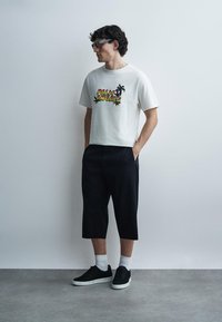 Άνδρας με λευκό μπλουζάκι Palm Angels, μαύρο κοντό παντελόνι, λευκές κάλτσες, μαύρα αθλητικά παπούτσια και γυαλιά, στέκεται μπροστά σε έναν απλό φωτεινό τοίχο.