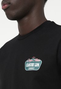 Črna bombažna majica s kratkimi rokavi, ki ima teal in belo logotipno oznako s rdečimi detajli. Okrugli ovratnik in gladka tekstura.