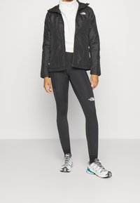 Svart isolerad jacka med huva, bärs över en vit polotröja, i kombination med svarta leggings och ljusgrå löparskor med detaljer.