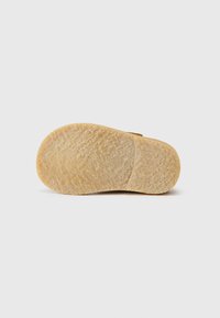 Suela de zapato de goma marrón claro con una superficie texturizada y forma redondeada, que presenta un emblema de marca visible y una ligera curvatura.