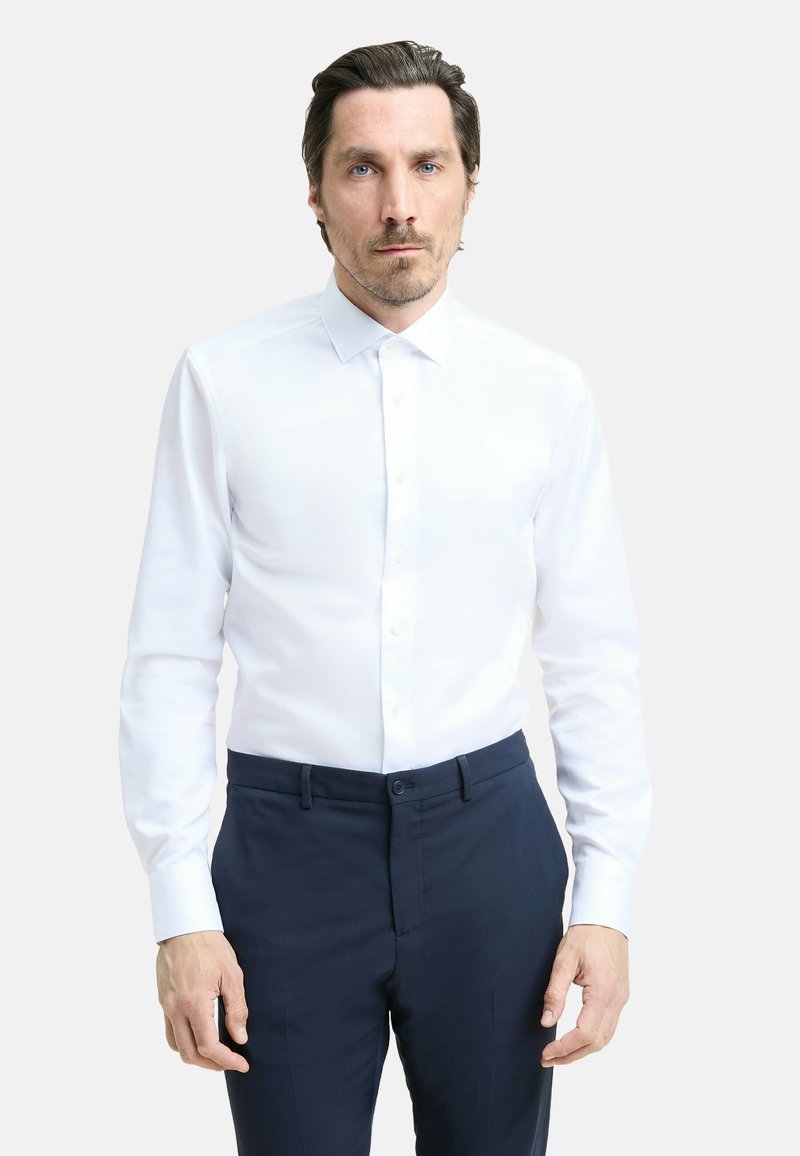 Camicia bianca con bottoni, maniche lunghe, colletto classico e texture liscia, abbinata a pantaloni eleganti blu scuro.