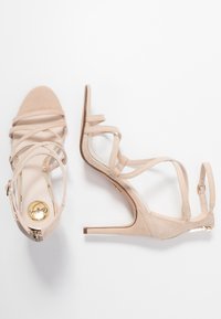 Beige sandaletter med hög klack i mockamaterial, öppen tå, flera remmar och en smal klack. Guldlogotyp på innersulan.