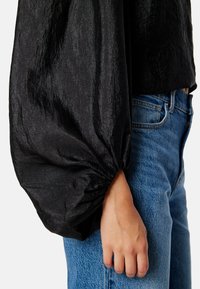 Zwarte blouse met textuur en volumineuze mouwen, met strakke manchetten, gecombineerd met blauwe denim jeans. Focus op de glans van de stof en de vorm van de mouwen.