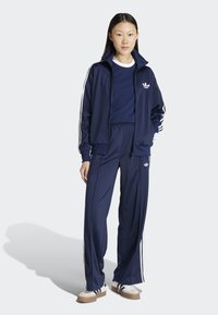 Marinblå träningsoverall med vita tre-stripes detaljer. Jacka med dragkedja, hög krage; byxor med elastisk midja. Modell som bär beiga sneakers.