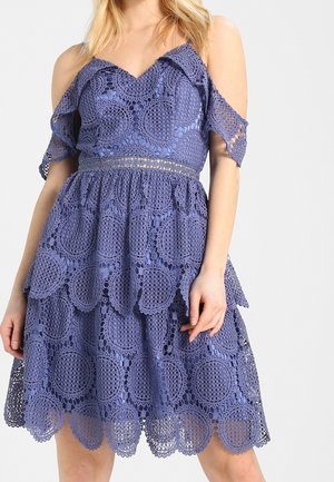 Femme portant une robe en crochet violette sans manches et épaules dénudées, ornée de motifs circulaires et d'un ourlet festonné.