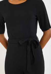 Černý žebrovaný overal s krátkými rukávy, přiléhavý střih a zavazovací pasový pásek. Materiál vypadá měkký s texturovaným povrchem.