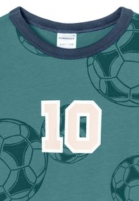 Groene katoenen T-shirt met een marineblauwe halslijn, groot "10" in lichtbeige en een achtergrondpatroon van voetbalballen in donkerder groene omlijningen.