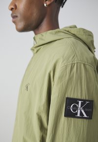 Giacca a vento verde oliva chiaro con una finitura testurizzata. Presenta una toppa nera con la scritta "Calvin Klein Jeans" e un piccolo logo sul davanti.