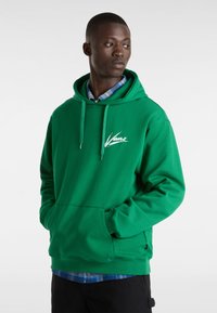Felpa verde realizzata in materiale morbido, con tasca frontale, coulisse regolabile e logo bianco sul petto. Indossata sopra una camicia a quadri blu.