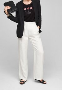 Blazer negro sobre una camisa oscura con diseño floral, combinado con pantalones blancos de pierna ancha y sandalias negras. Sosteniendo un bolso de mano, fondo neutro.