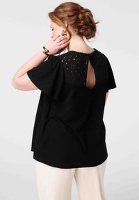 Haut noir à manches courtes avec un tissu texturé et un panneau en dentelle au dos avec une ouverture en forme de clé. Associé à un pantalon de couleur claire.