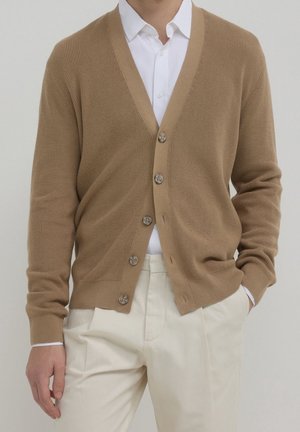 Mann trägt beige, geknöpfte Strickjacke über weißem Hemd mit Kragen und cremefarbenen Hosen, mit einer Hand in der Tasche vor einfachem Hintergrund.