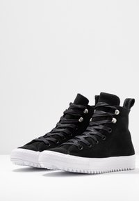 Schwarze Wildleder-High-Top-Sneakers mit silbernen Ösen und schwarzen Schnürsenkeln. Weiße Gummisohle mit einem Zickzack-Muster. Einfaches, modernes Design.