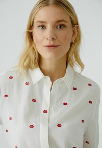 White button-up shirt featuring red embroidered lip motifs. The fabric appears smooth with a slight sheen, and has a classic collar and buttons.