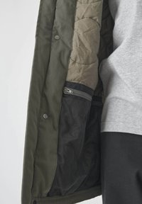 Grüner Jackeninnenraum mit gestepptem, hellkhakifarbenem Futter und einer schwarzen Netzinnentasche. Verfügt über Druckknöpfe und einen sichtbaren Reißverschluss.