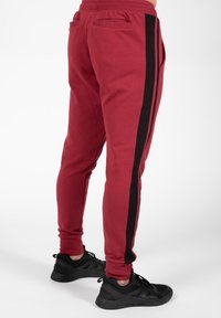 Rode joggingbroek met zwarte zijkanten, taps toelopende pasvorm, elastische tailleband en twee achterzakken. Draagt met zwarte sportschoenen.