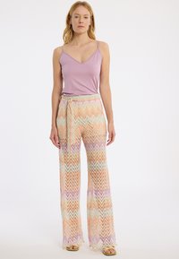 Fliederfarbenes Spaghettiträgershirt und weit geschnittene Hose mit Zickzackmuster in Pastellfarben und Bindegürtel. Glatte Textur und leichtes Material.