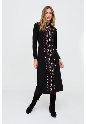 Frau steht und trägt ein schwarzes langärmliges Kleid mit vorne mehrfarbigem Halbmondmuster, schwarze Stiefel und burgunderrote Strumpfhosen vor weißem Hintergrund.