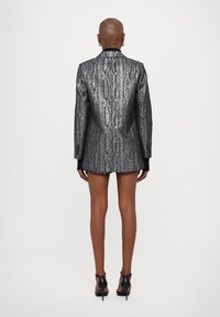 Strieborný metalický blazer so vzorom hadej kože, so štruktúrovanými plecami, dlhými rukávmi a priliehavým strihom, skombinovaný s čiernymi podpätkami.