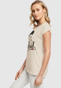 Hellbeige T-Shirt mit einem grafischen Druck, der ein Porträt einer Frau und das Wort "Rebel" zeigt. Kurzärmlig, lockere Passform, weicher Baumwollstoff.