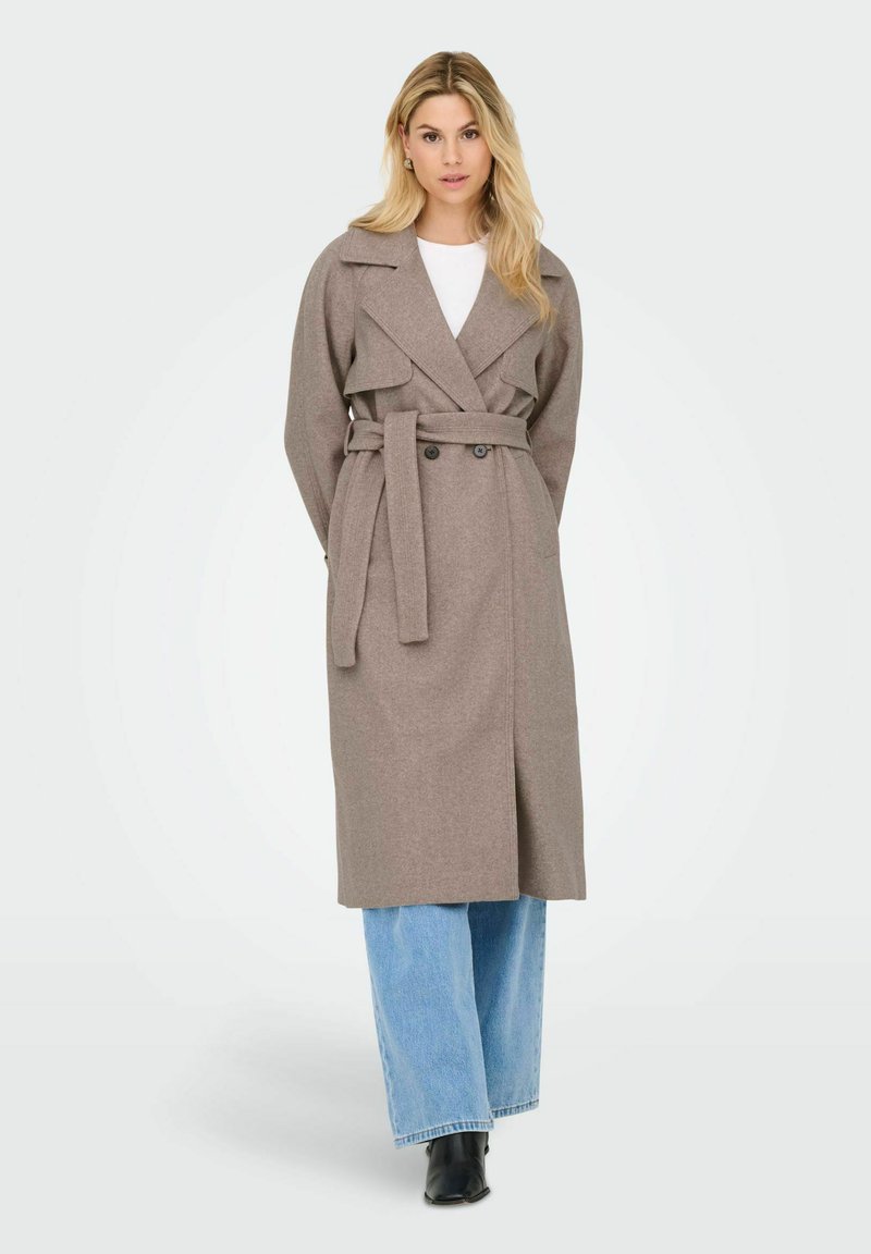 Beige bælte trenchcoat med brede reverser og to knapper, parret med lyseblå vide bukser og sorte ankelstøvler.