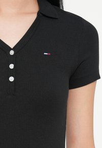 Juodas ribotas polo marškinėliai su V formos iškirpte, trumpomis rankovėmis, penkiomis sagomis ir mažuoju siuvinėtu logotipu raudona, balta ir mėlyna spalvomis krūtinėje.
