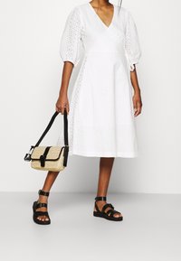 Bílé bavlněné šaty s dírkovaným vzorem, výstřihem do V a nadýchanými rukávy. Doplněné texturovanou béžovou kabelkou přes rameno a černými sandály s pásky.